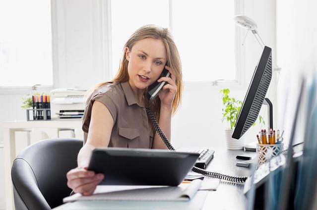 Eine Frau sitzt im Büro am Schreibtisch und schaut auf ein Tablet während sie telefoniert.